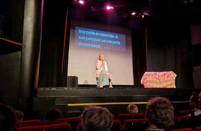 Mathilde Cabanis parle sur une scène de théâtre, derrière elle s'affiche un écran de sous-titrage